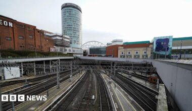 A view from above of train tracks coming in and out of Birmingham New Street. The tracks are empty with no passengers on the platforms. A large cylinder-shaped building is seen in the distance with other retail and leisure buildings around the station.