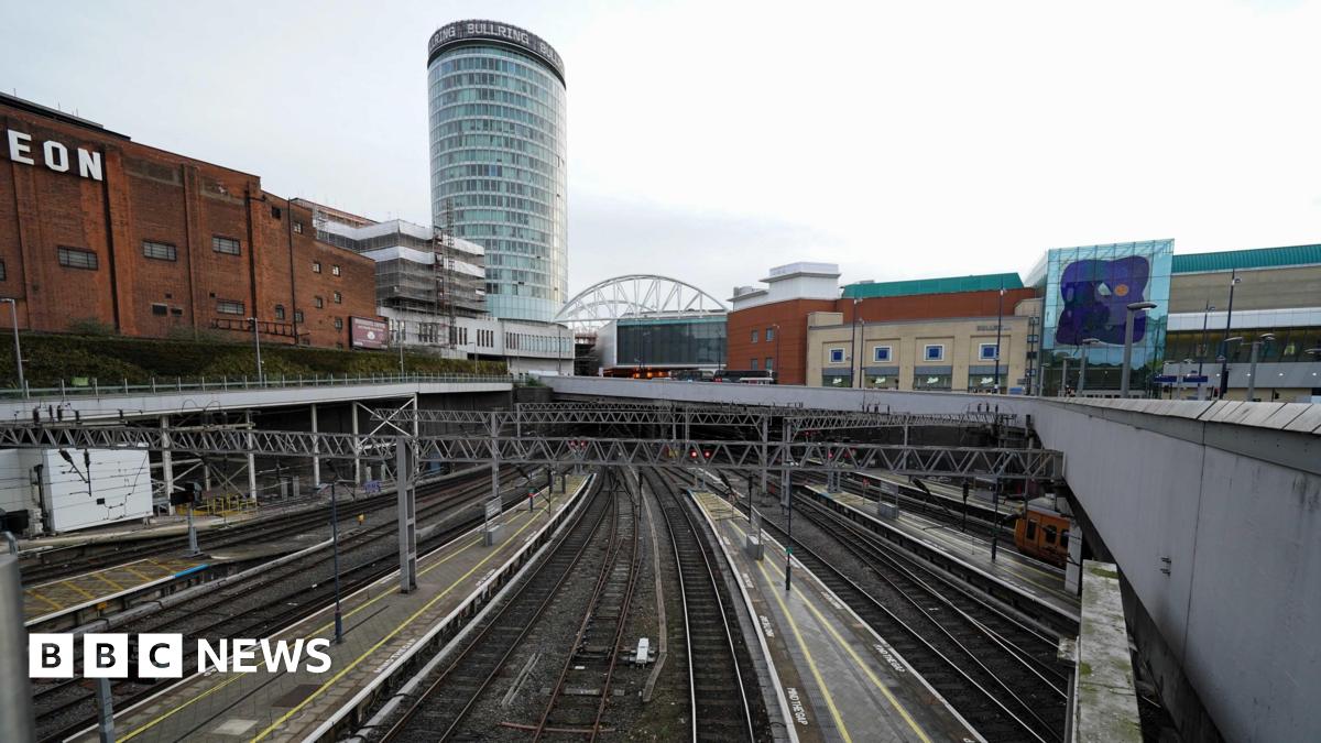 A view from above of train tracks coming in and out of Birmingham New Street. The tracks are empty with no passengers on the platforms. A large cylinder-shaped building is seen in the distance with other retail and leisure buildings around the station.