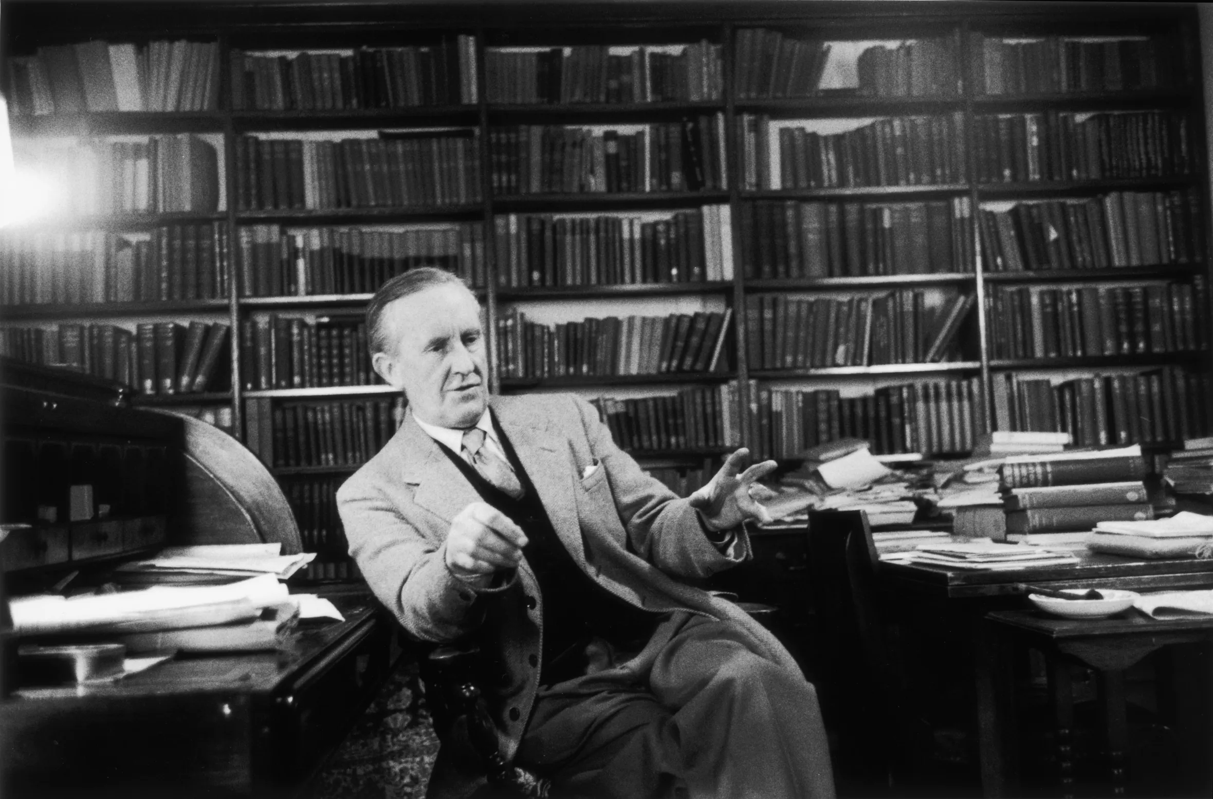 J.R.R. Tolkien, author of 'The Hobbit' and 'The Lord of the Rings,' gestures while seated at a desk in a library filled with books.