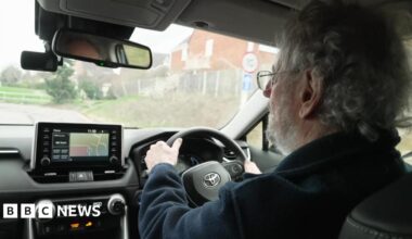 Photograph of Turner driving, taken over his shoulder. You can see the back of Turner's head - he has short, curly grey hair and a beard. He is wearing a pair of thin-framed glasses, and a dark blue fleece. The interior of his car is black, with a silver Toyota logo on the wheel. Outside the windscreen, you can see Turner is driving down a residential street.