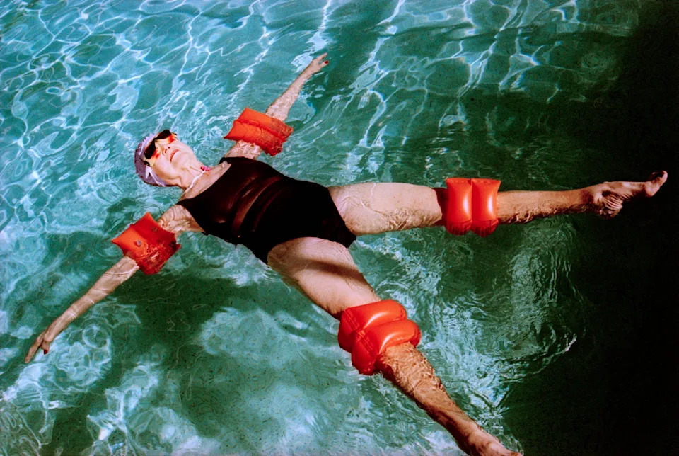 Person floating in a pool wearing a black swimsuit and orange floaties on arms and legs, appearing relaxed and peaceful