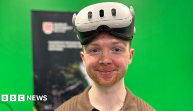 A man with red facial hair and wearing a white VR headset smiles at the camera. He is wearing a brown shirt. He is standing in front of a Queen's University Belfast banner and a green screen.