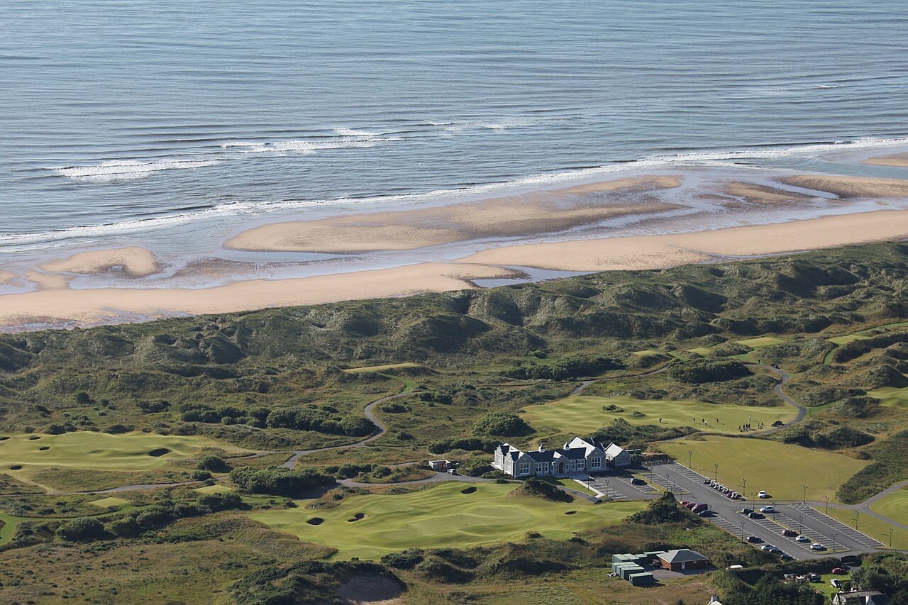 Aerial view of a coastal golf course with a clubhouse, car park and manicured greens set among sand dunes beside the sea.