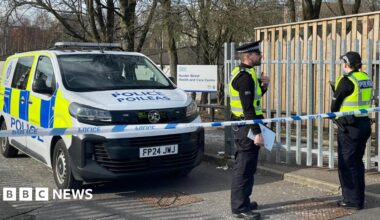Two police officers stand behind a line of police tape beside a police van. In the background is an NHS sign saying "Hunter Street Health and Care Centre".