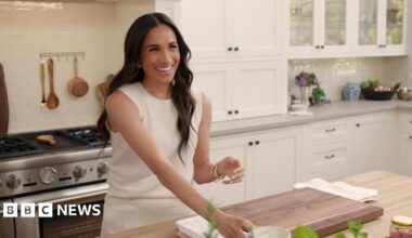 The Duchess of Sussex in a kitchen smiling and wearing a white outfit