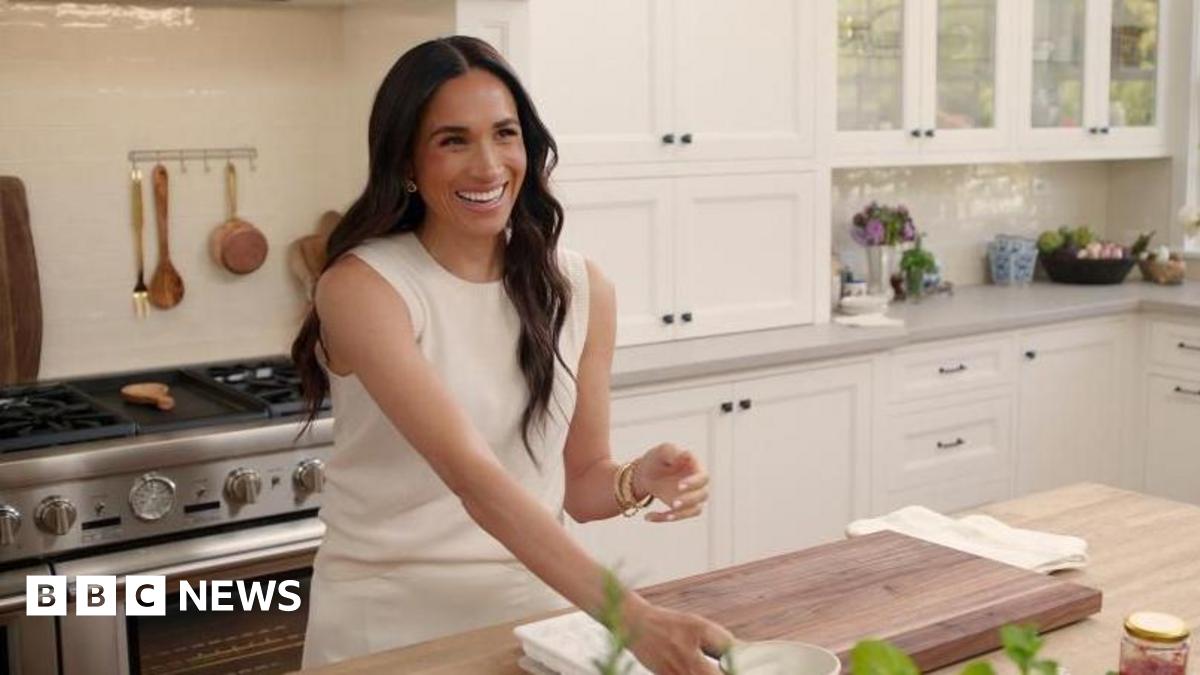 The Duchess of Sussex in a kitchen smiling and wearing a white outfit