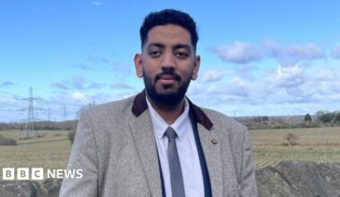 A man, wearing a light grey blazer with dark coloured collar over a white shirt and blue tie, looks at the camera. There is a stone wall behind him, as well as a field with trees and bushes.