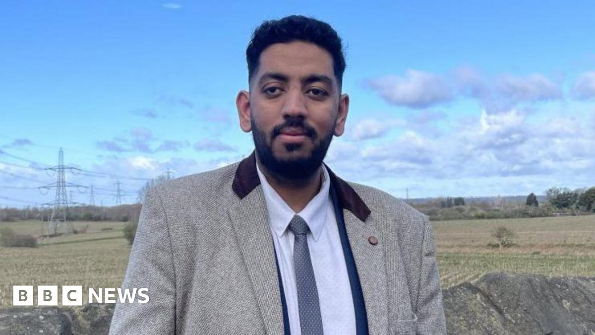 A man, wearing a light grey blazer with dark coloured collar over a white shirt and blue tie, looks at the camera. There is a stone wall behind him, as well as a field with trees and bushes.