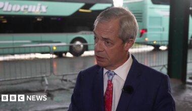 A man with grey hair, wearing a dark blue suit, white shirt and red tie. There are two green buses stationed behind him.