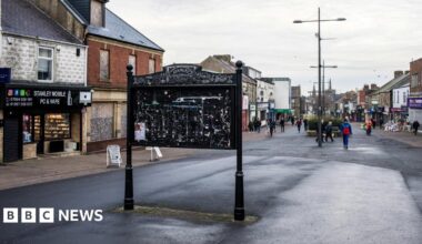 Stanley High Street. A large black notice board stands in the middle of the street. It is empty. A number of vacant and boarded up shops line the street.