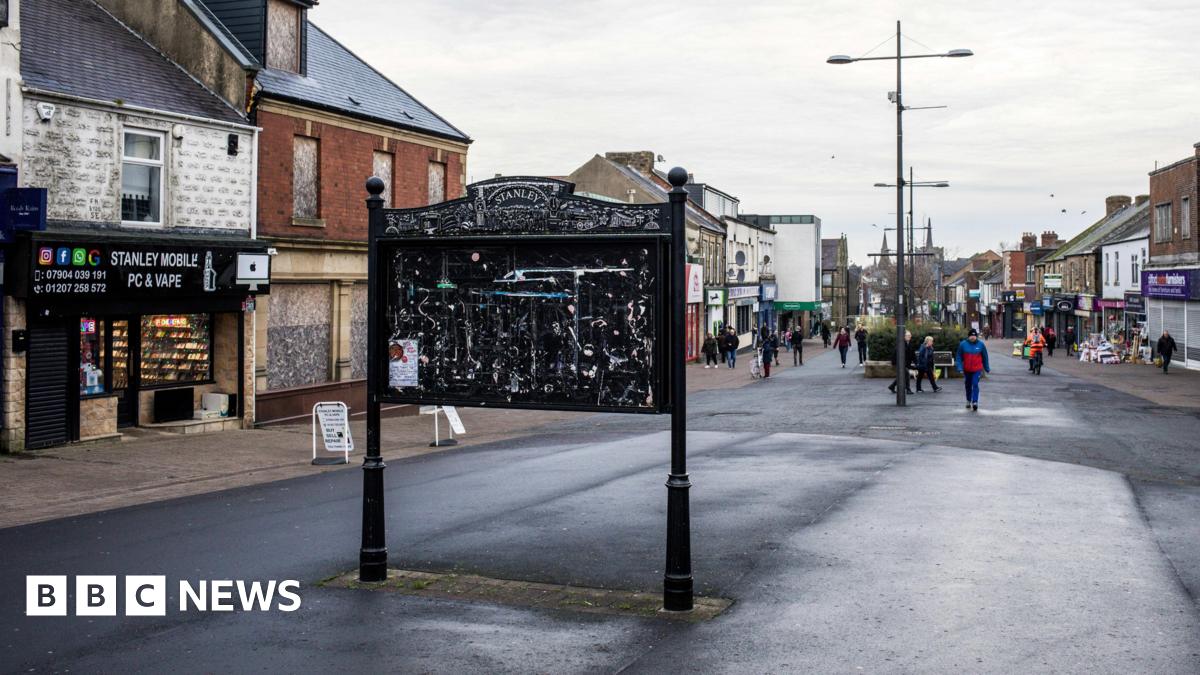 Stanley High Street. A large black notice board stands in the middle of the street. It is empty. A number of vacant and boarded up shops line the street.