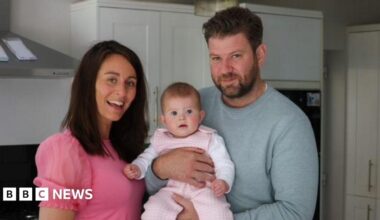 Man and woman smile widely with a baby in the man's arms.