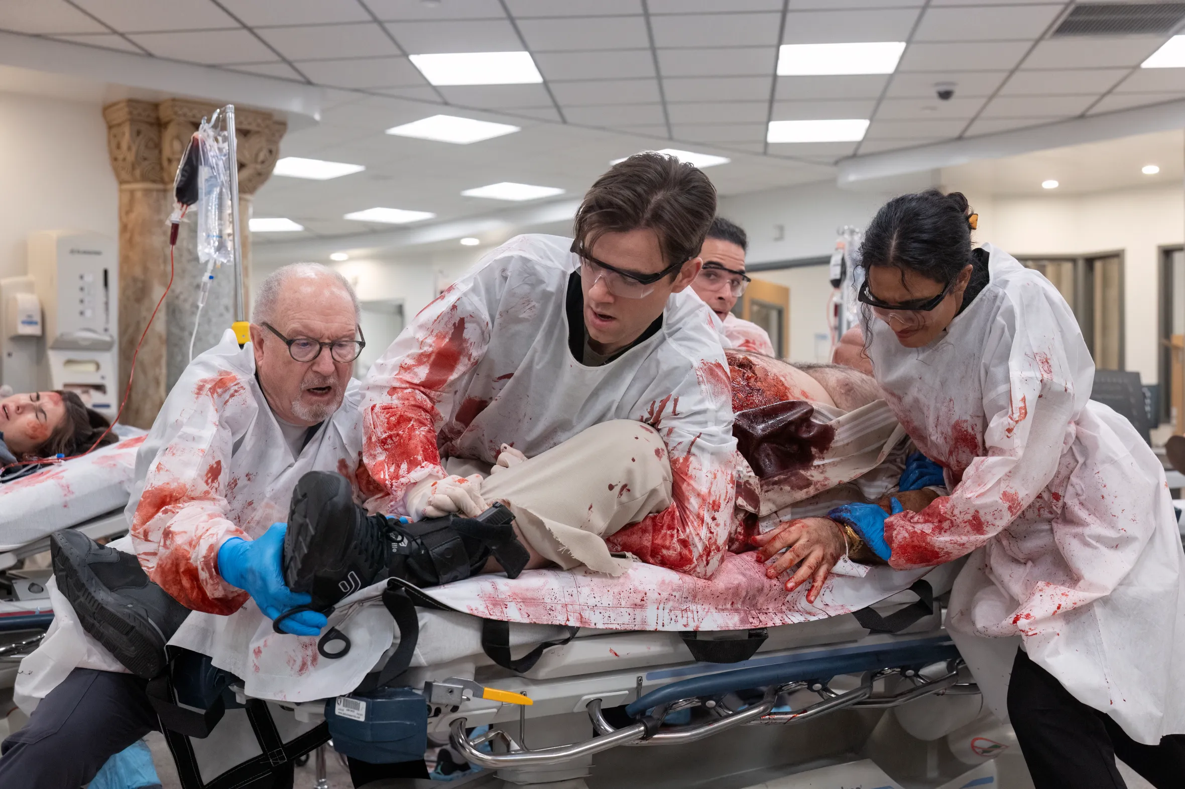 Doctors treat a severely wounded patient with blood all over their surgical gowns and the stretcher, while another patient is visible in the background.