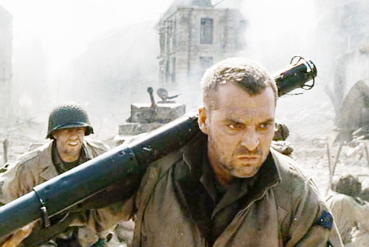 An individual is seen carrying a substantial weapon while navigating through a chaotic, smoke-filled environment. In the background, remnants of a destroyed building and the presence of armored vehicles suggest a military conflict setting. Another soldier, equipped with a helmet and protective gear, follows closely behind, contributing to the scene's intensity.