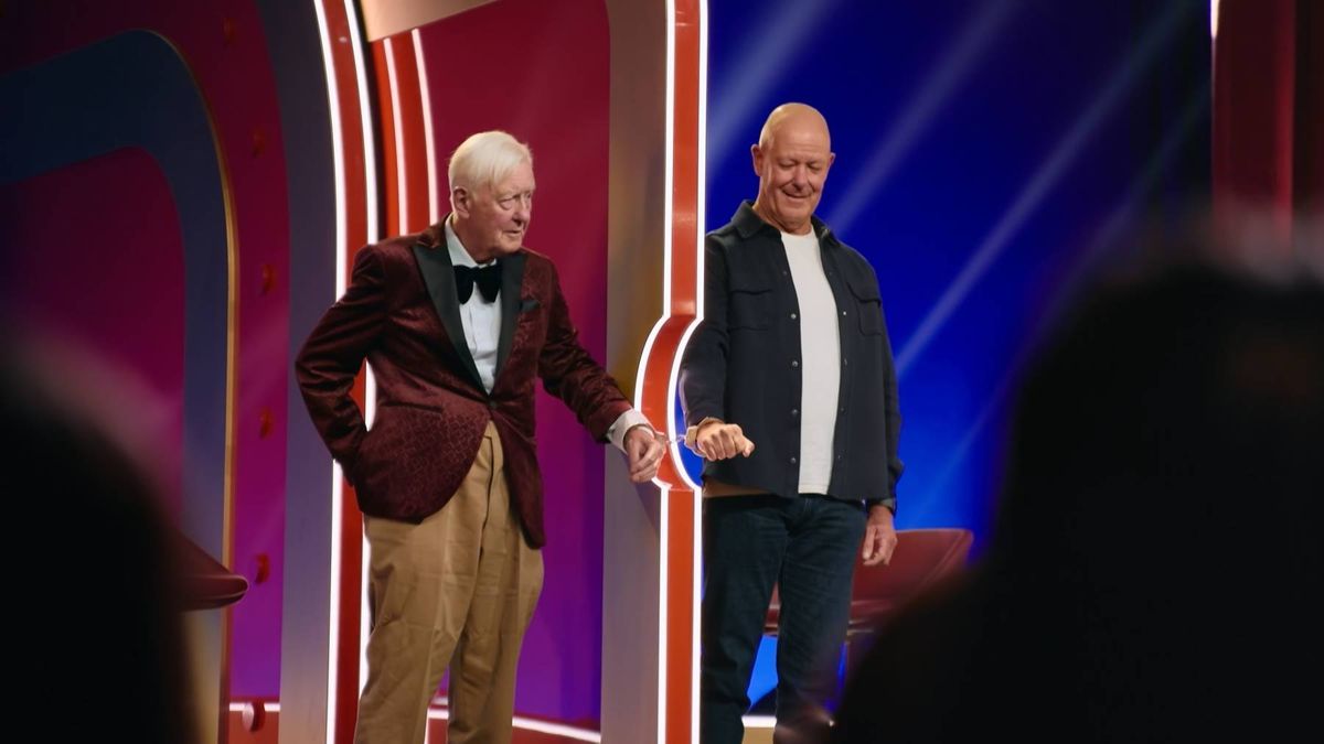 Two individuals are standing on a stage adorned with vibrant red and blue backdrops. One person is dressed formally in a dark suit and bow tie, while the other is wearing a black jacket. They appear to be engaged in conversation or interaction, with the formal attire suggesting a significant event or presentation.