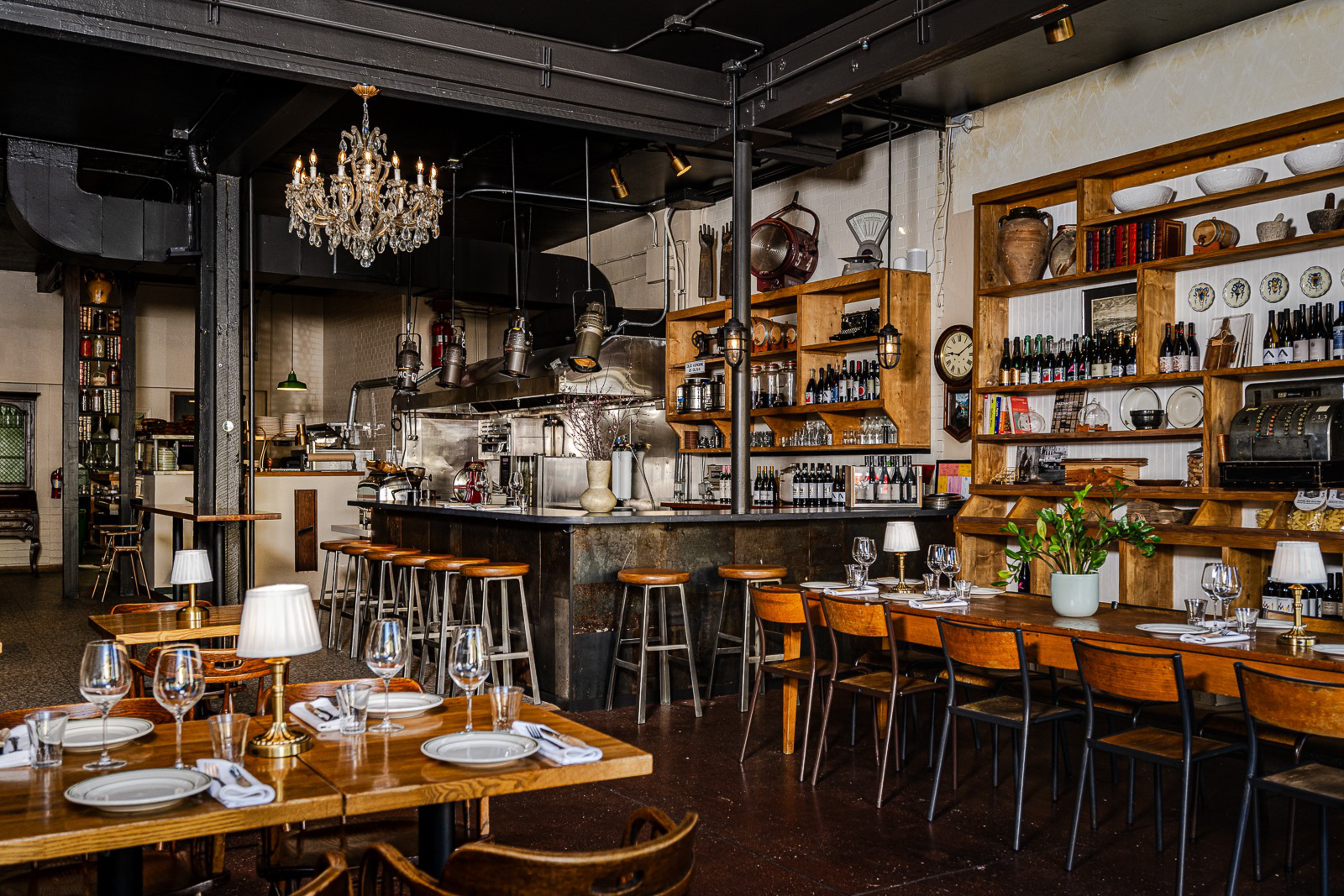 Wood-finished tables and chairs, wine bottles, décor, crystal chandelier, and a bar counter with stools create a cozy, rustic restaurant interior.