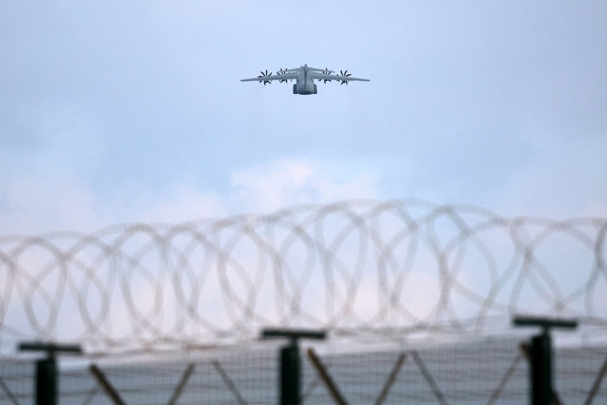 A large plane in the sky, seen through the perspective of someone behind a barbed-wire fence.