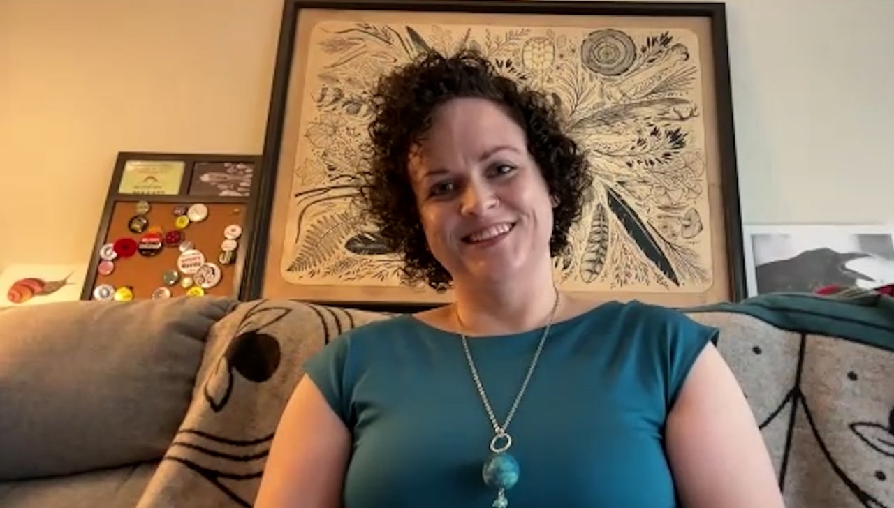 A woman with short, curly dark hair wearing a blue short-sleeve top and necklace. 