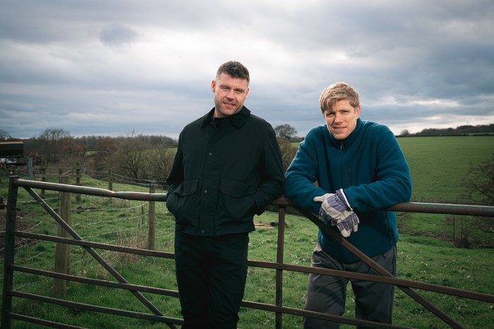 Aaron and Robert stood against a fence on open farmland in a scene from Emmerdale
