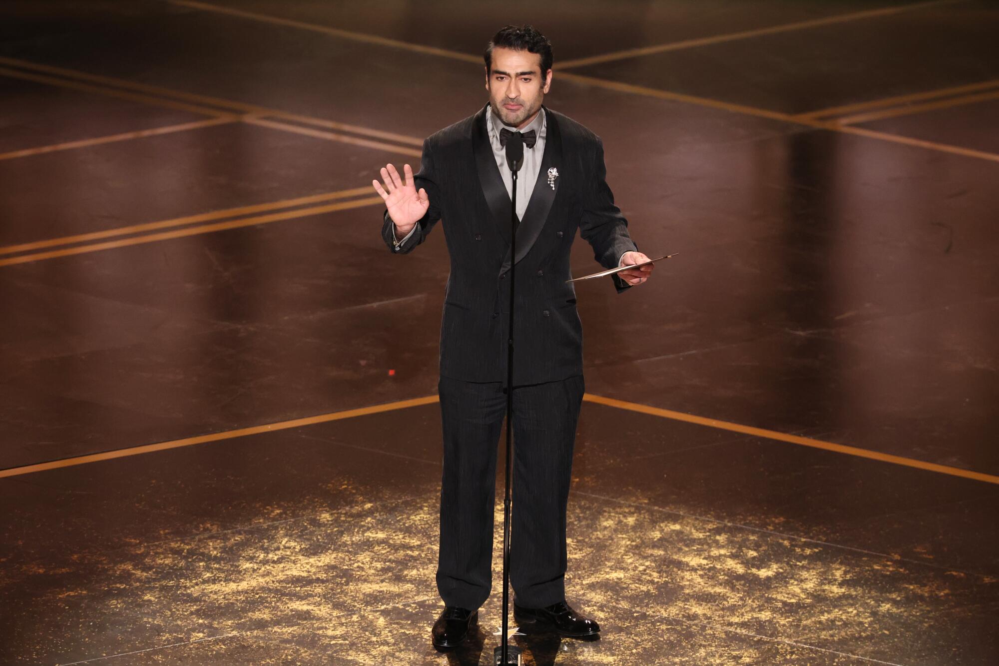 Kumail Nanjiani on stage in a tux holding up his palm 