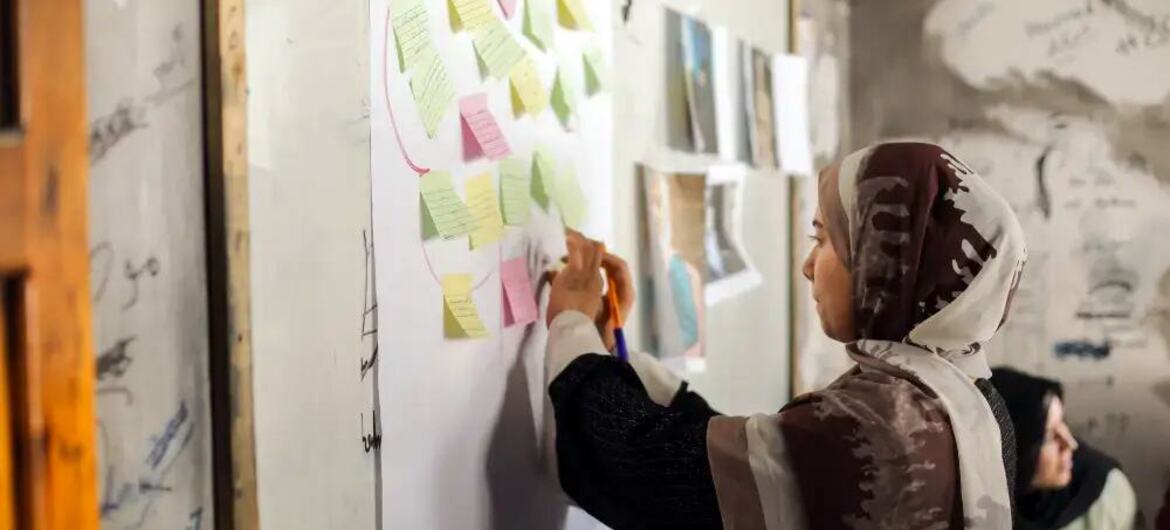 A woman wearing a headscarf actively participates in a planning session, writing on a whiteboard covered with colourful sticky notes.
