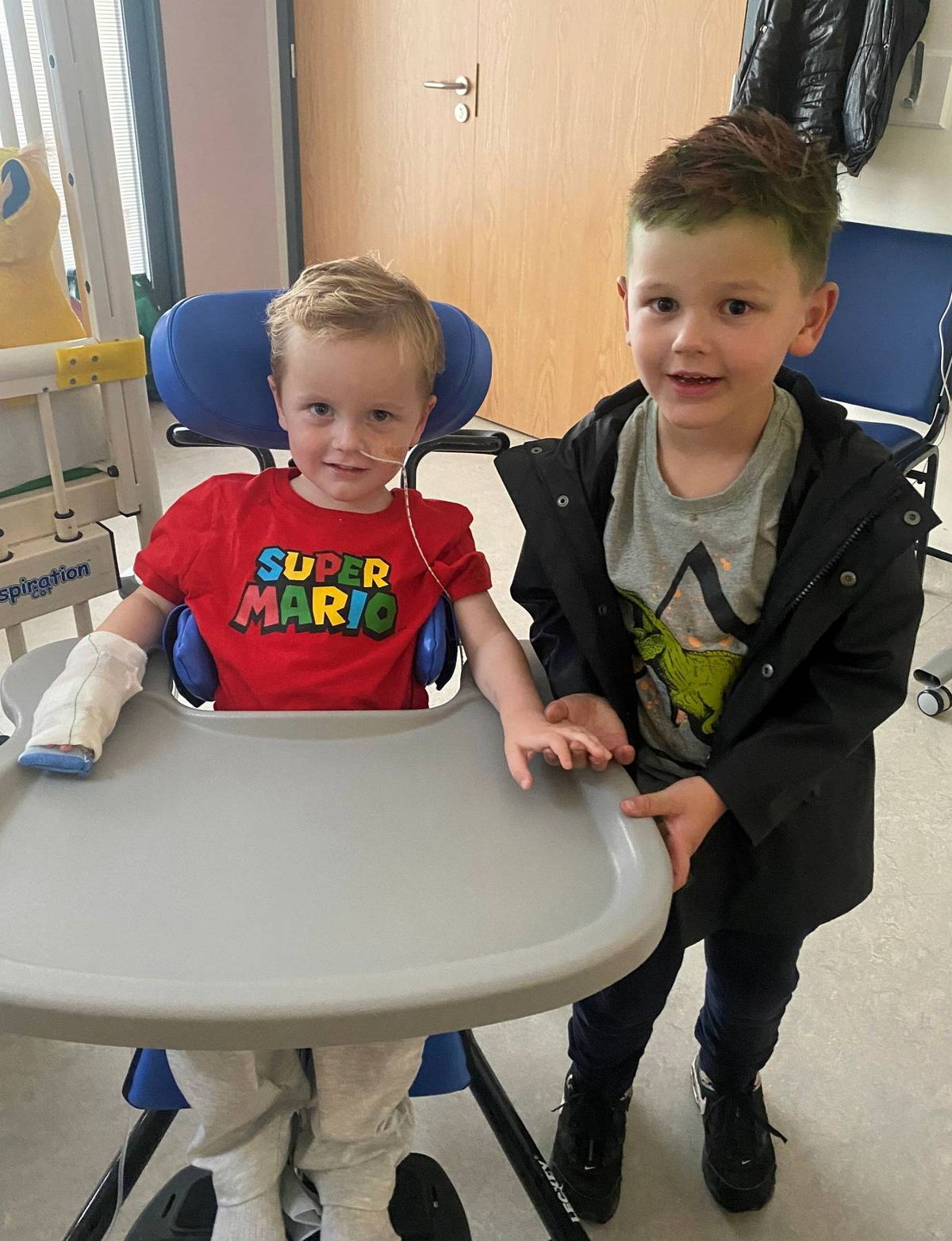 Two young boys are sitting together in a hospital room. One boy is seated in a blue chair with a white arm brace, while the other boy stands next to him, smiling and looking towards the camera. The setting appears to be a hospital room, indicated by the medical equipment and the hospital chairs in the background.