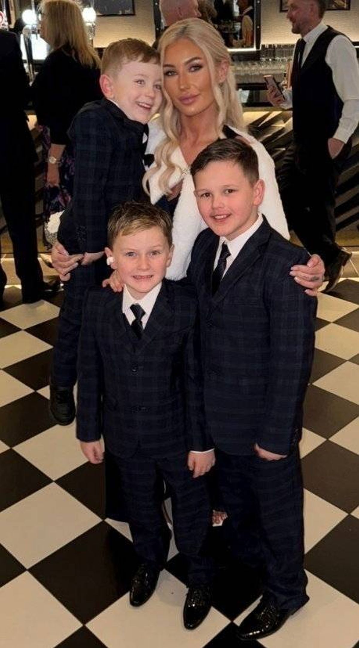 A family consisting of a woman and two young boys dressed in formal attire, smiling and posing for a photograph, with a blurred background of people and an indoor setting.