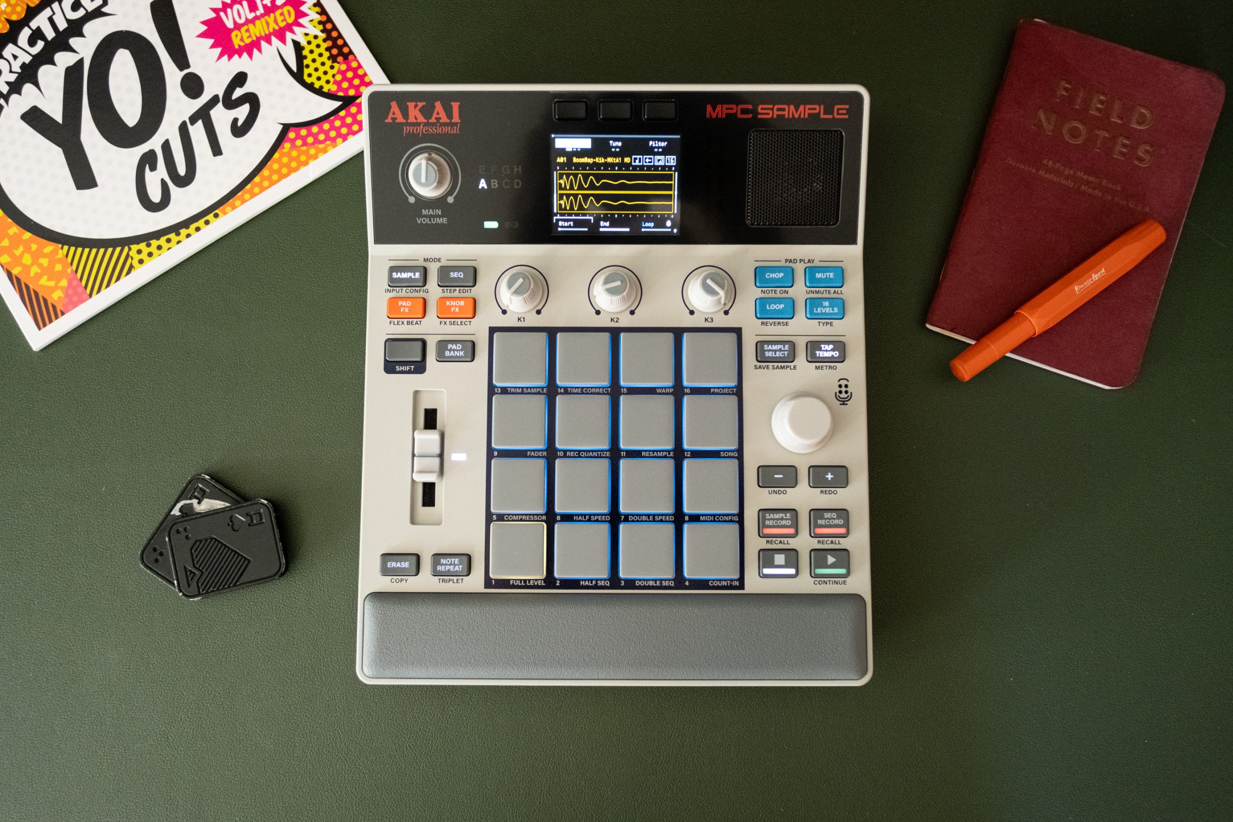 The MPC Sample on a desk with a scratch vinyl, fidget toy, pen, and a very pretty Field Notes notebook.