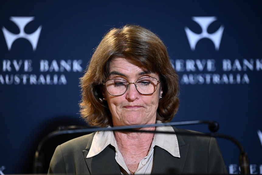 A woman with short brown hair and glasses looking down at her notes behind a lectern with two microphones.