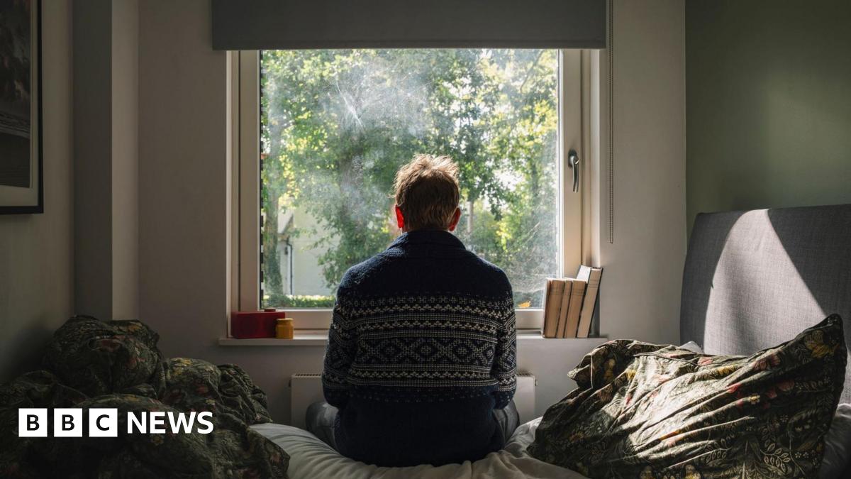 A man is seen from behind as he is sat on a bed looking out onto a sunny day.