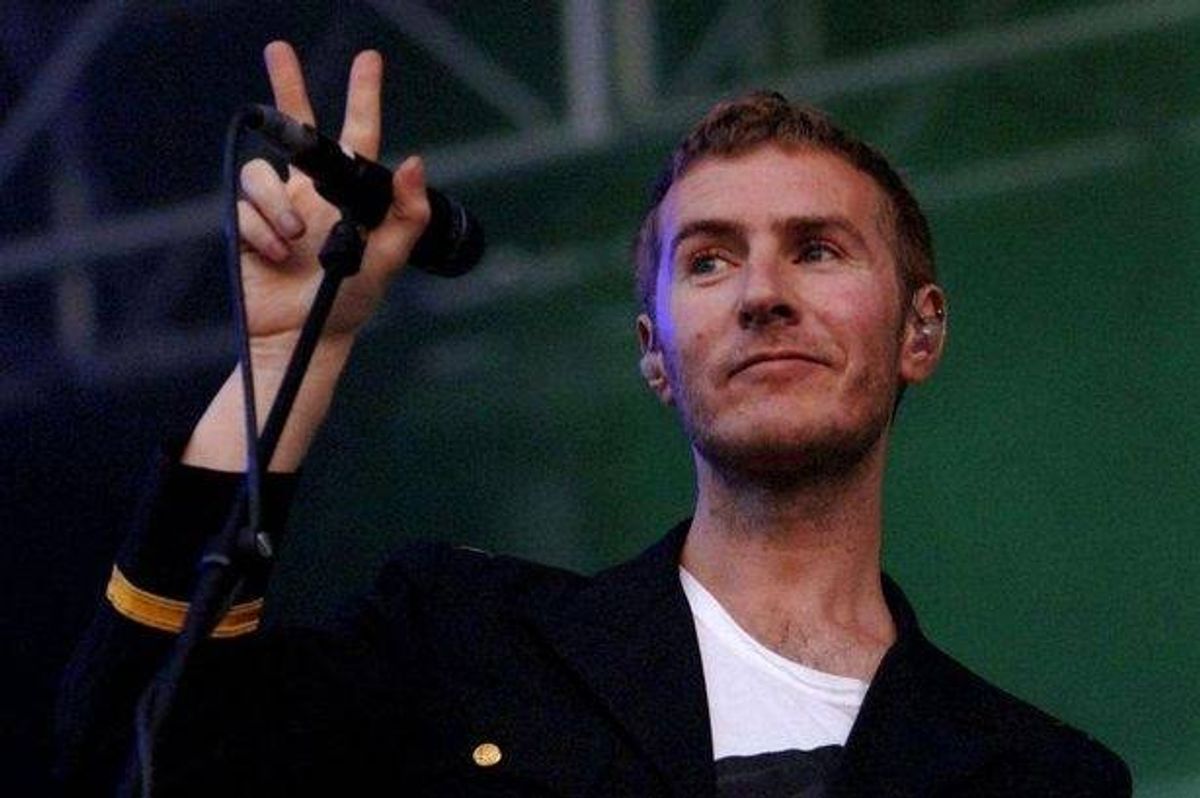 An individual stands on a stage, gesturing with one hand while holding a microphone in the other. He is dressed in a dark jacket over a white inner shirt, with a backdrop featuring a vibrant green hue and architectural elements.