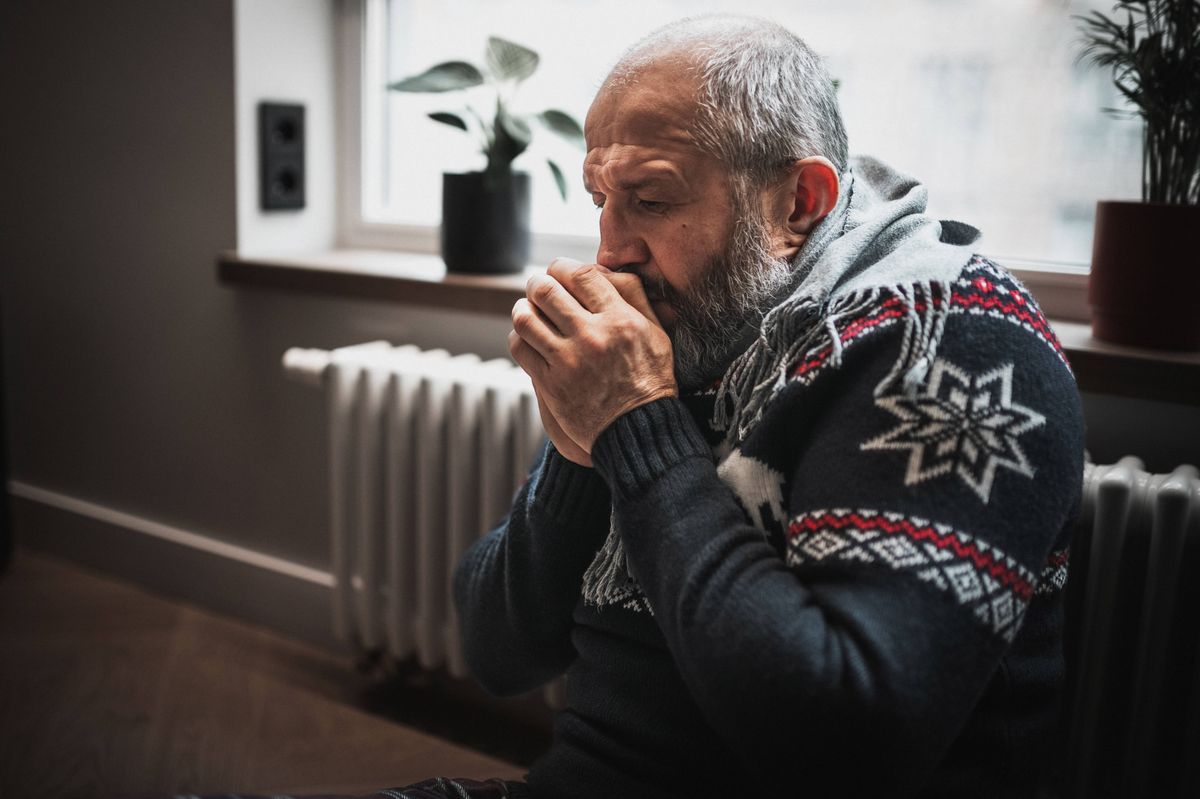 Unwell man feeling cold in home. He's trying to stay warm next to the radiator.