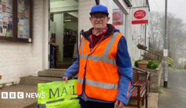 A man in his 70s wearing glasses, a blue cap and blue jacket with a flourescent orange high-vis vest. In his right hand he is holding his flourescent yellow paperbag with 'Mail' printed on it in capital letters. He is standing outside a whitewashed post office.