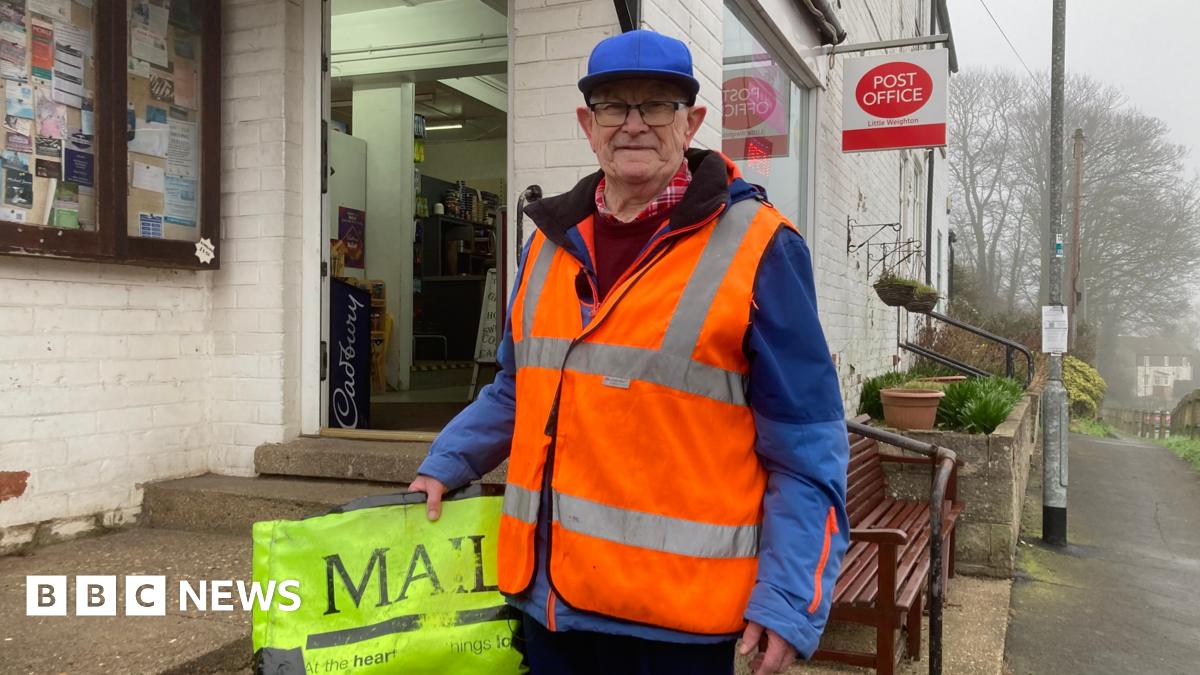 A man in his 70s wearing glasses, a blue cap and blue jacket with a flourescent orange high-vis vest. In his right hand he is holding his flourescent yellow paperbag with 'Mail' printed on it in capital letters. He is standing outside a whitewashed post office.