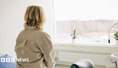 A stock image of a blonde woman sitting on a bed in a hospital room facing towards a window. She has wavy blonde hair and a heavy khaki shirt. There is a pillow covered with a blue pillowcase visible on the bed and to the right of the frame a black stool can be seen.