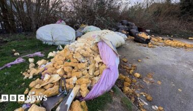 Several large bags of waste dumped on a supermarket car park. One of the bags coloured purple has split and what is believed to be foam loft installation has spilled out of it among other waste. The bags of rubbish are in front of trees.