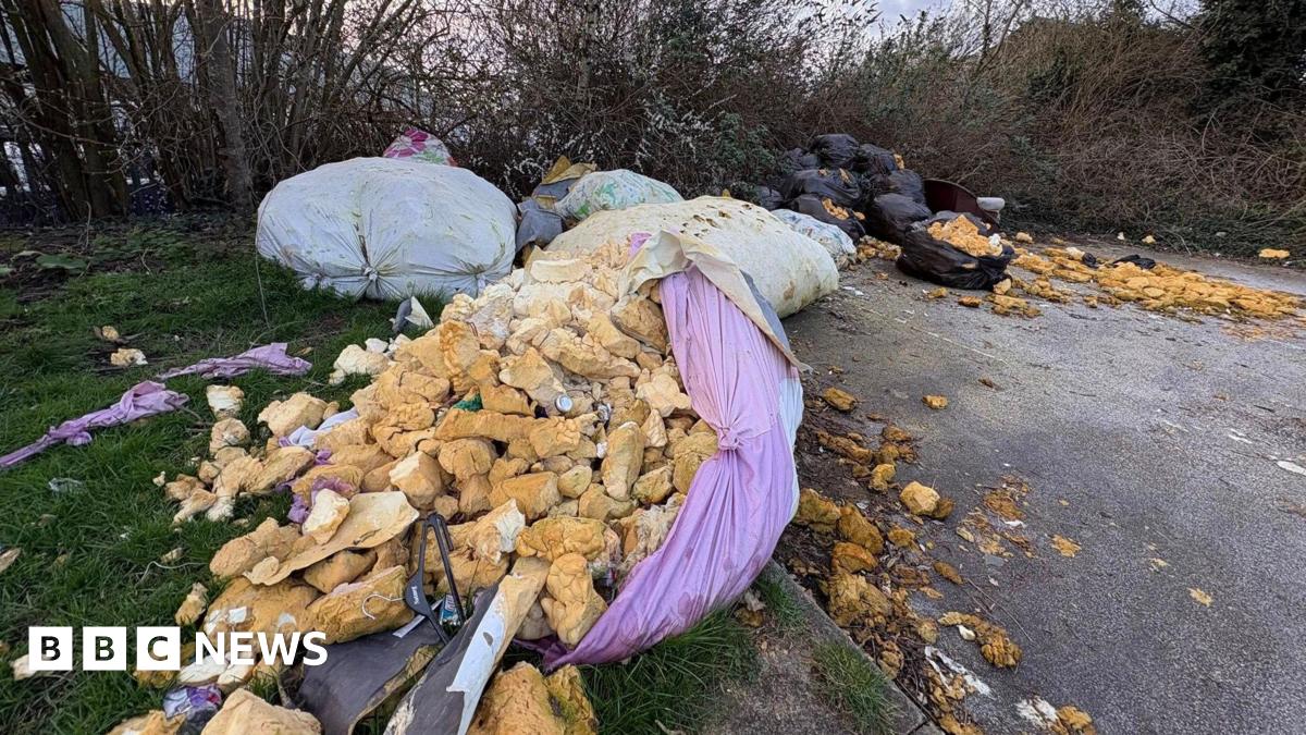 Several large bags of waste dumped on a supermarket car park. One of the bags coloured purple has split and what is believed to be foam loft installation has spilled out of it among other waste. The bags of rubbish are in front of trees.