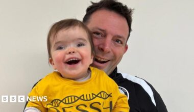 A man in a black tracksuit top holds up his young daughter who is wearing a yellow T shirt and is smiling broadly
