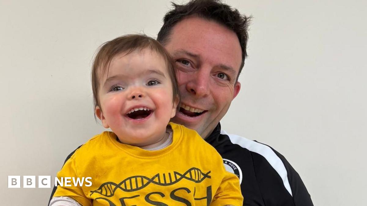 A man in a black tracksuit top holds up his young daughter who is wearing a yellow T shirt and is smiling broadly