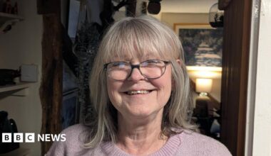 Jacky Smith smiles at the camera inside her home. She has short grey hair and wears glasses along with a light pink knitted jumper.