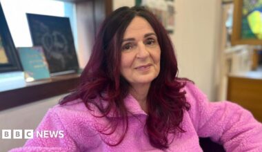 Trish Prosser has long dark red hair and looking at the camera as she wears a pink fleece. Behind her on a recessed windowsill are pieces of artwork. Next to the window is a cream wall.