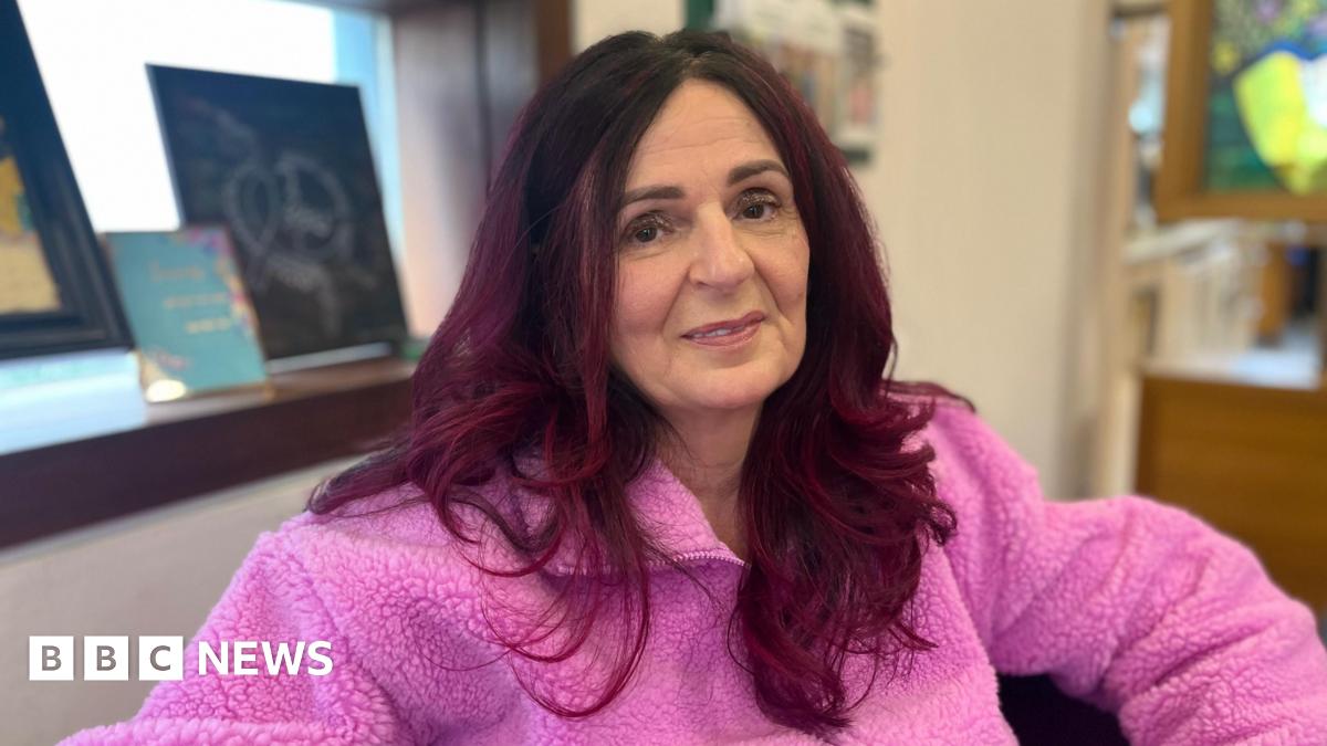 Trish Prosser has long dark red hair and looking at the camera as she wears a pink fleece. Behind her on a recessed windowsill are pieces of artwork. Next to the window is a cream wall.