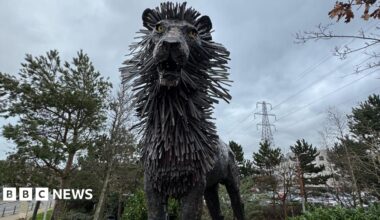 A statue of the Gruffalo , a mythical creature with a large head, orange eyes, a wide month, a round torso, horns, tusks and large claws.  The statue is standing in a wooded area with one paw leaning on a wooden post.