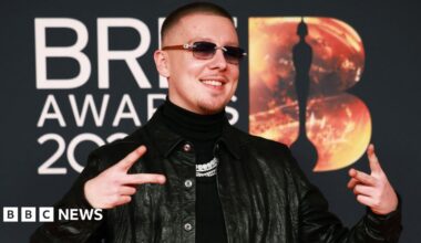 Aitch, with short brown and dark shades wearing a dark polo neck swearer and black leather jacket poses on the red carpet during the BRIT Awards at the Co-op Live Arena, in Manchester, Britain, 28  February  2026.
