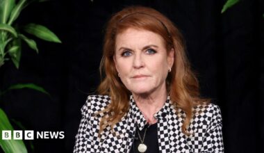 Sarah Ferguson, the then Duchess of York, attends the traditional Easter Sunday Mattins Service at St George's Chapel, Windsor Castle on April 20, 2025. She has long red hair with a black hairband and is dressed in a white jacket with back trim and is wearing black gloves. One arm is raised as she waves.
