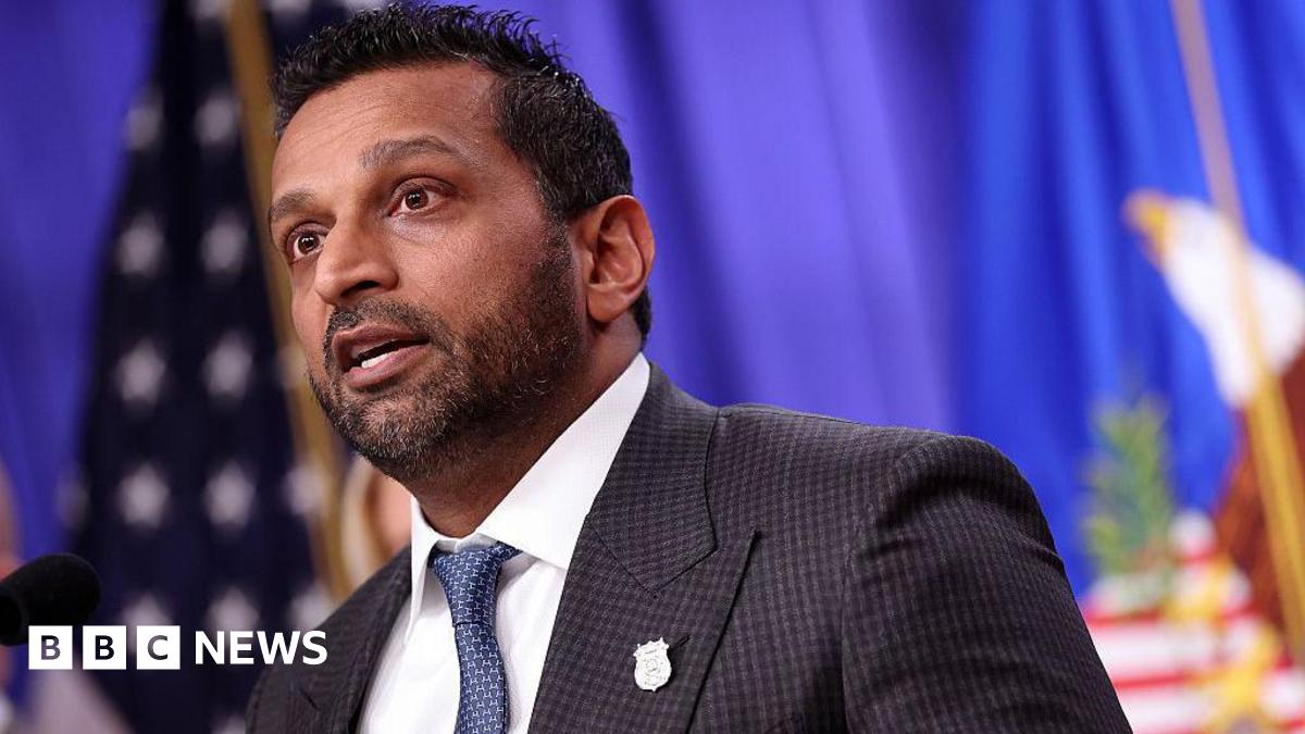 close-up of Kash Patel speaking, wearing a suit and tie in front of American flag