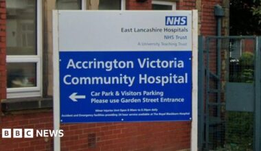 A street view image of Accrington Victoria Hospital, a red brick building with blue and white signs either side of a white awning with blue legs over the entrance and a man walking under it to the front door