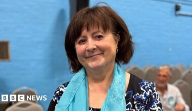 A woman wearing a blue scarf is standing in a sports hall with a blue wall. There are chairs and other people behind her. She is smiling for the photograph.