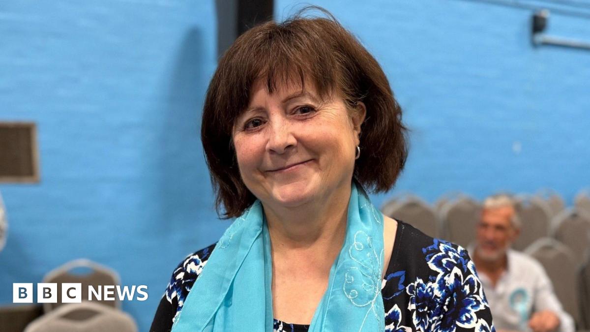 A woman wearing a blue scarf is standing in a sports hall with a blue wall. There are chairs and other people behind her. She is smiling for the photograph.