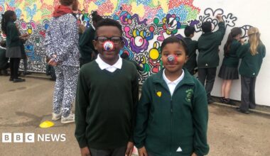 A man with red hair and beard is dressed in a black and white top, featuring cartoon drawings of a variety of characters. He is wearing one red nose with black and white doodle illustrations, and four of his fingers with a red nose with black and white doodle illustrations.